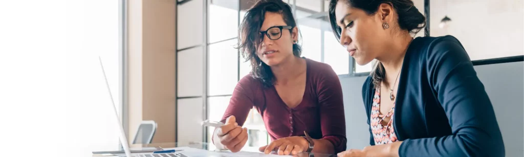 Two colleagues sit at a desk in an office, discussing a project.